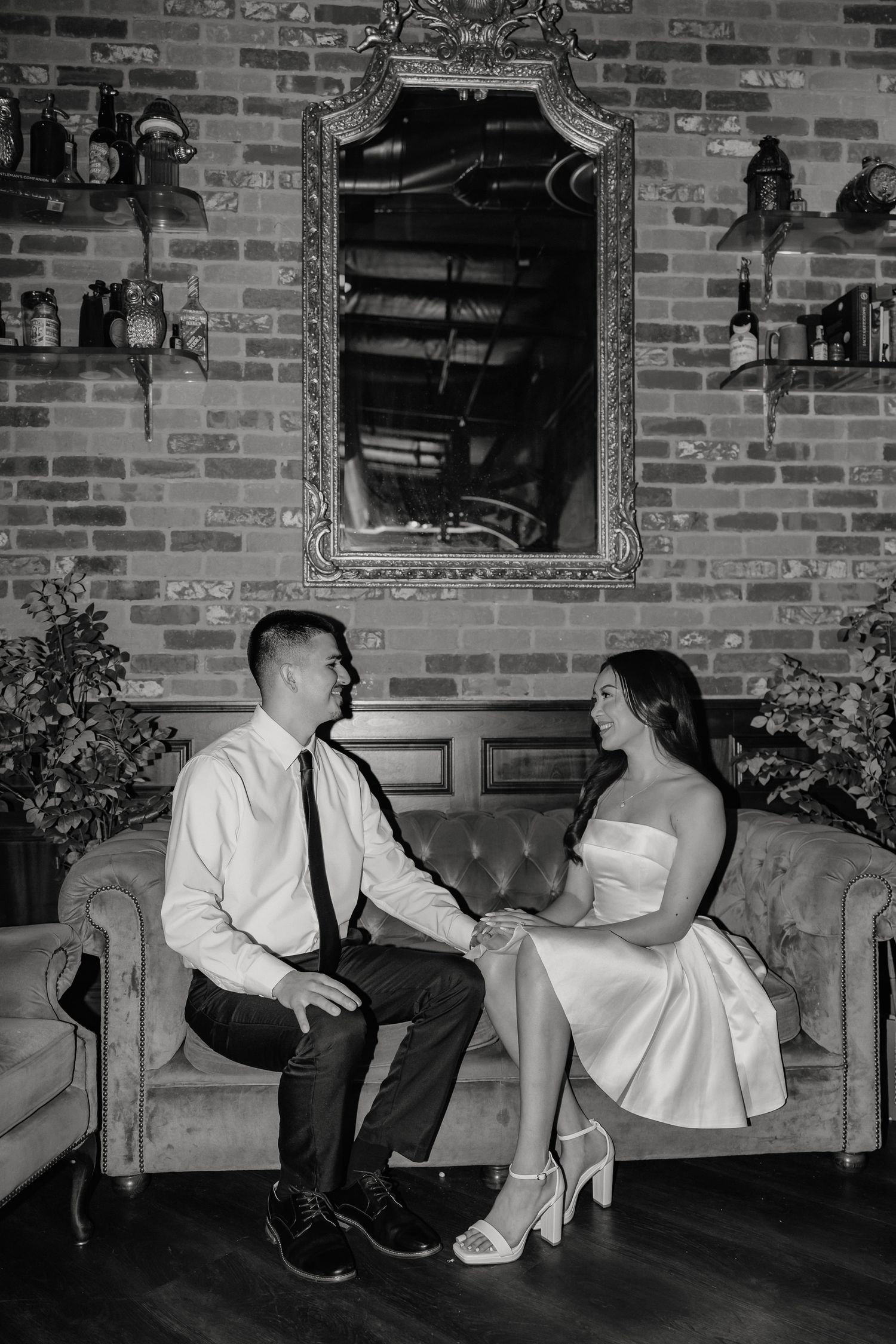 A couple sits together on a vintage couch in black and white, with an ornate mirror on the brick wall behind them.