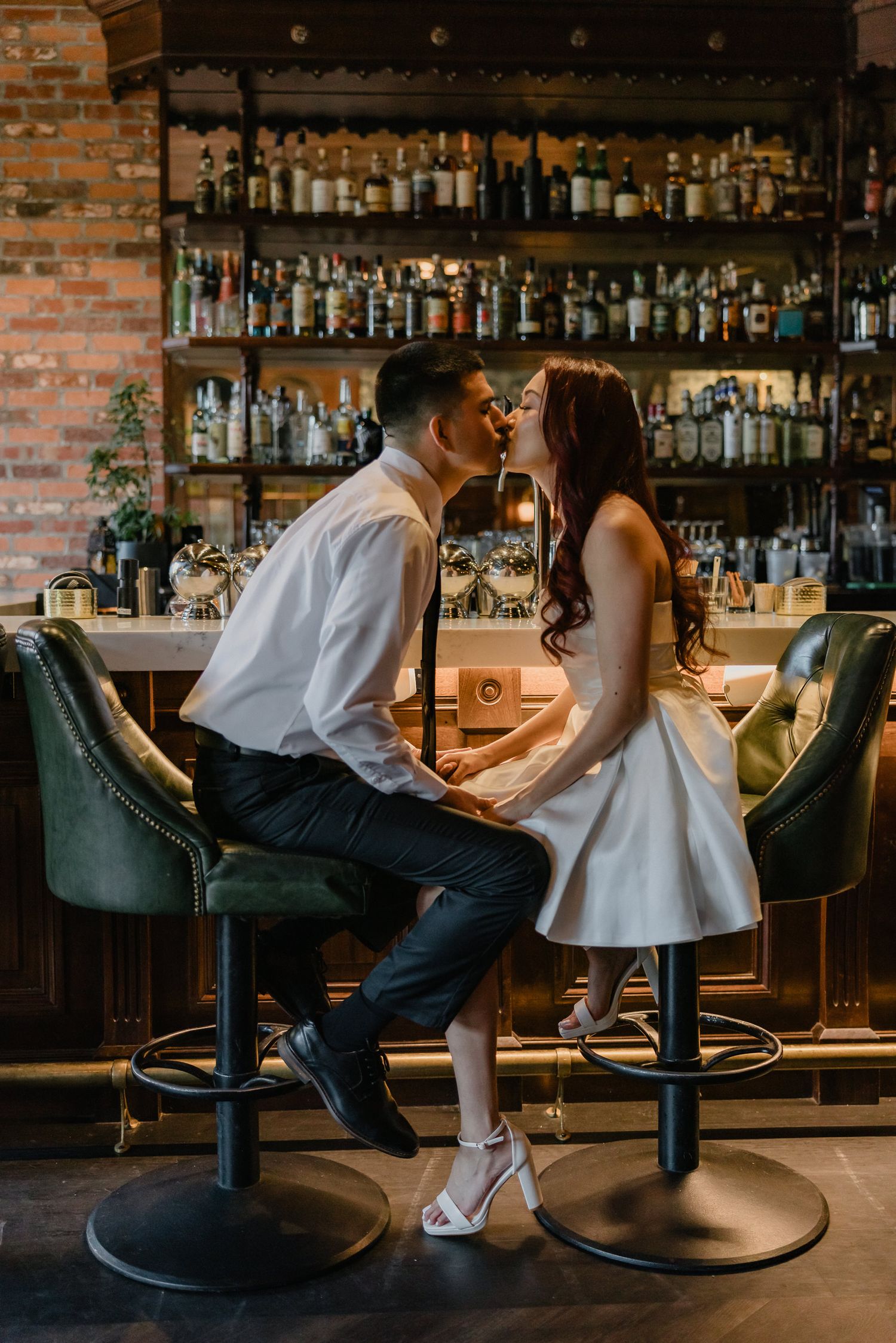 Romantic couple sharing an intimate moment at a stylish bar with vintage brick walls and shelves of liquor bottles.