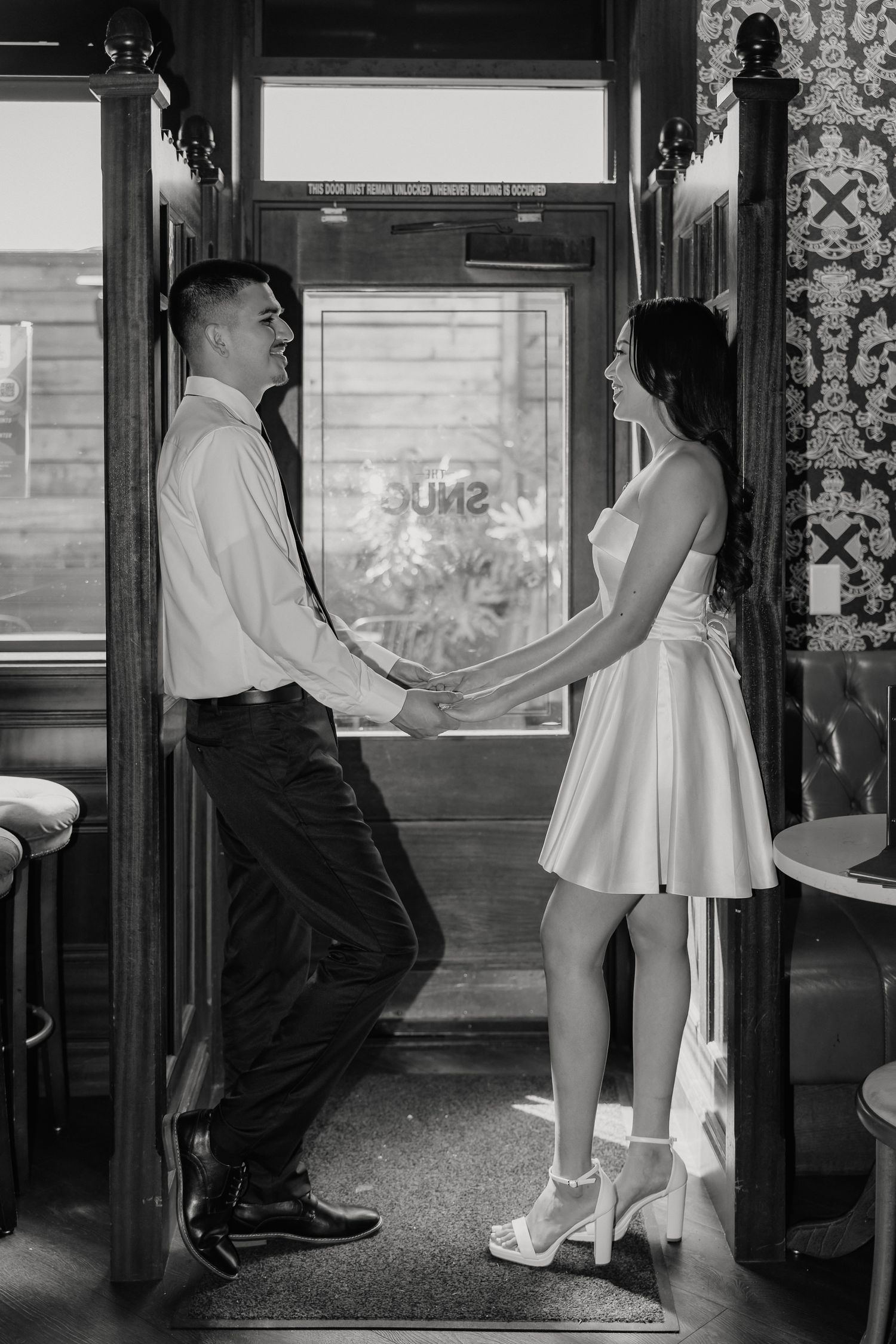 A couple in vintage-style clothing hold hands while standing in a train car doorway in black and white photography.