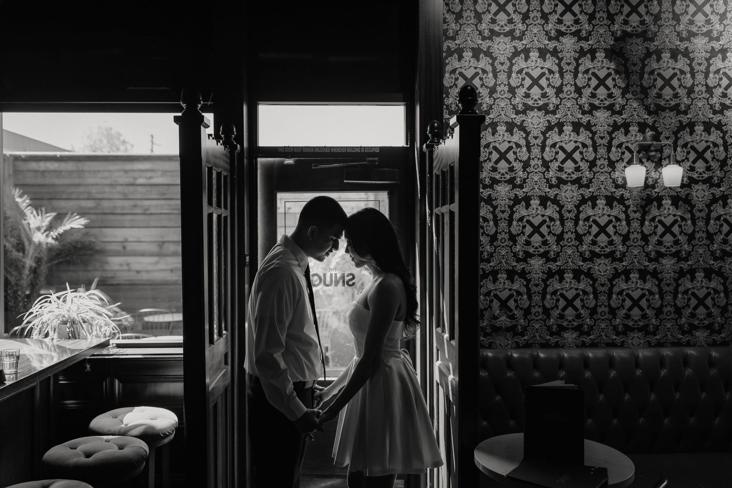 A romantic silhouette of a couple embracing in a doorway against ornate wallpaper in black and white photography.
