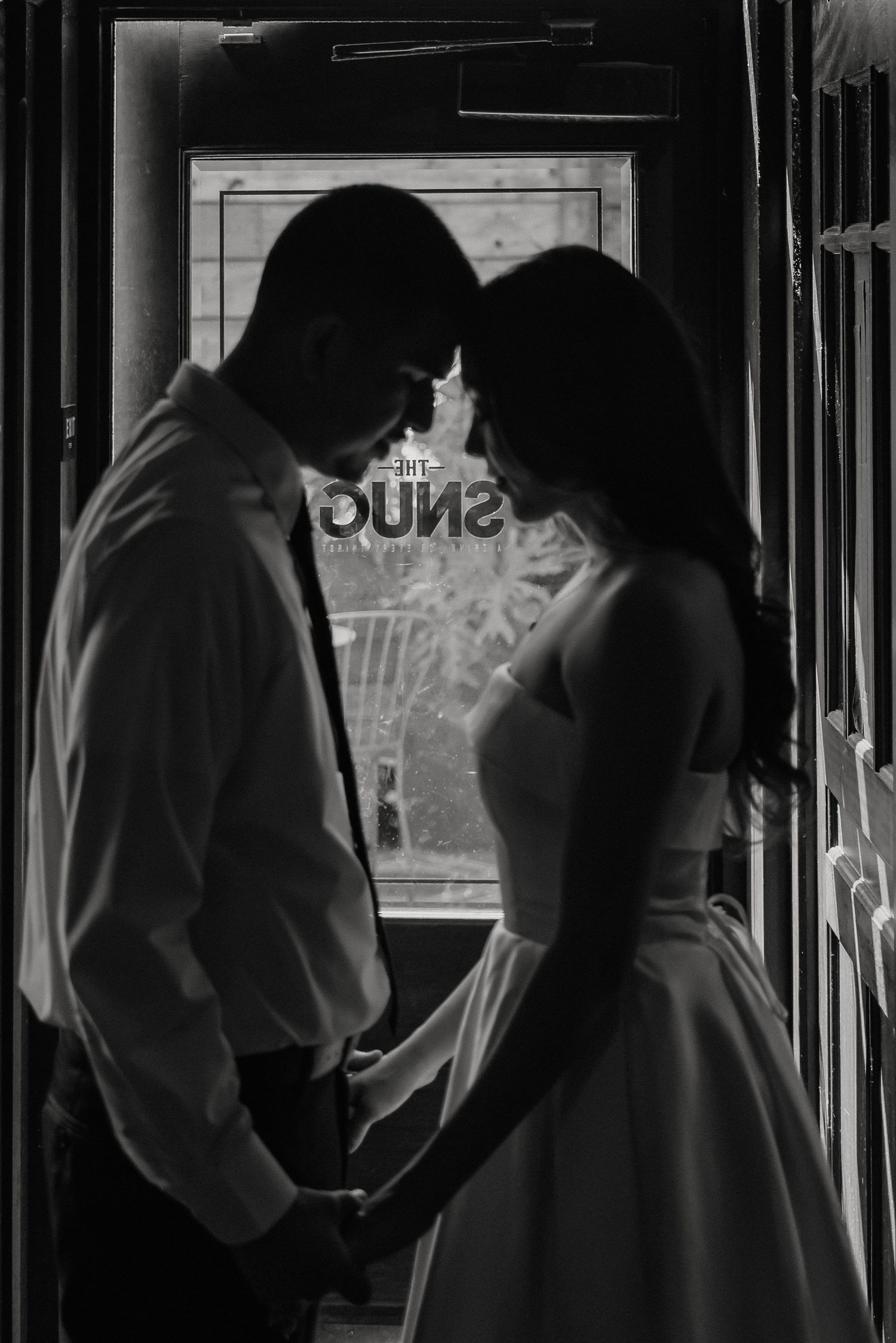 A silhouetted couple stands intimately in a doorway, their dark figures framed by natural light through a window.