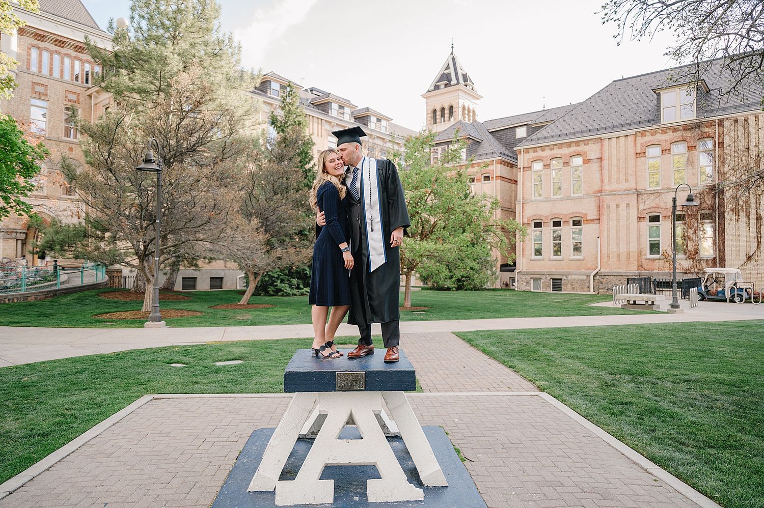 Two graduates celebrate their commencement standing on a large UA logo emblem on campus at the University of Arkansas.