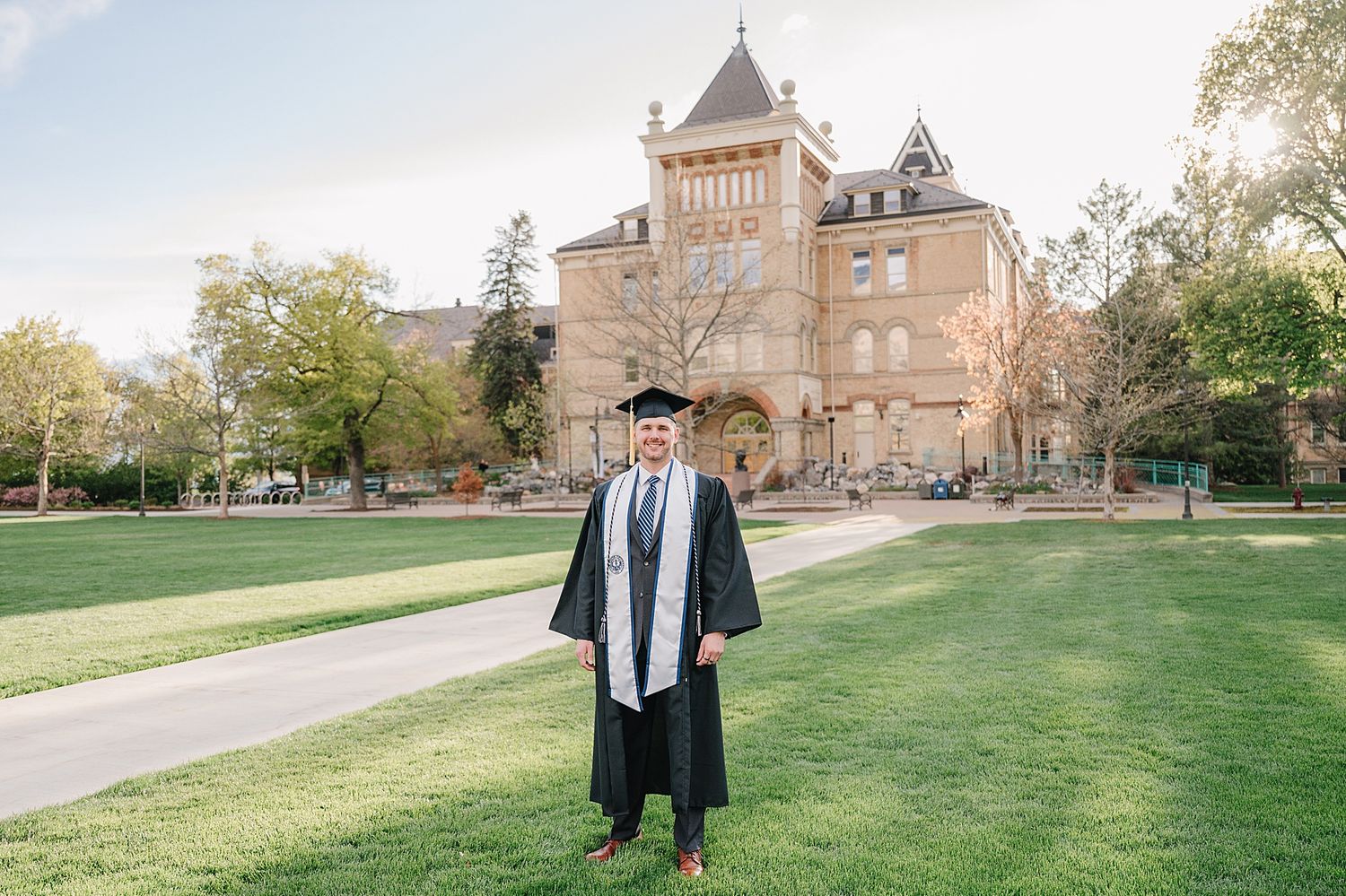Graduate in traditional cap and gown posing in front of historic collegiate building on sunny day.