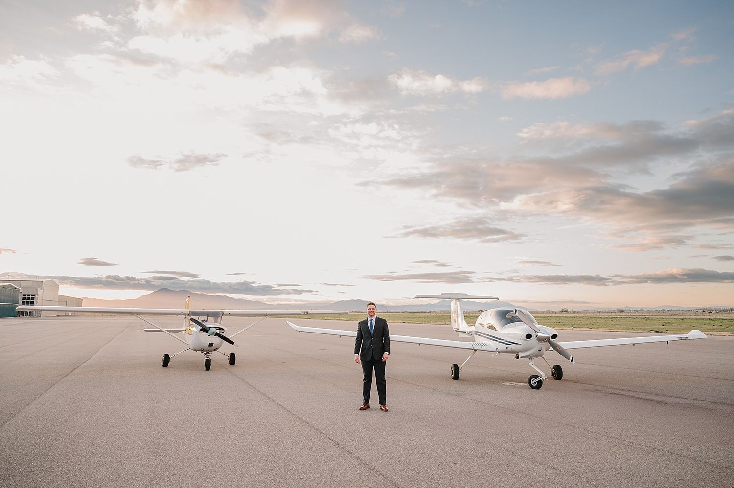 A person stands between two small aircraft on a tarmac during sunset with cloudy skies overhead.