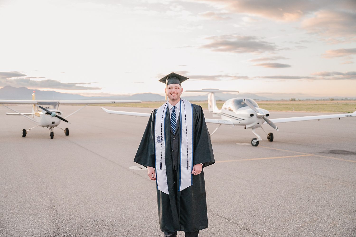 Graduate in cap and gown standing on airport runway with two small aircraft parked in background during sunset.