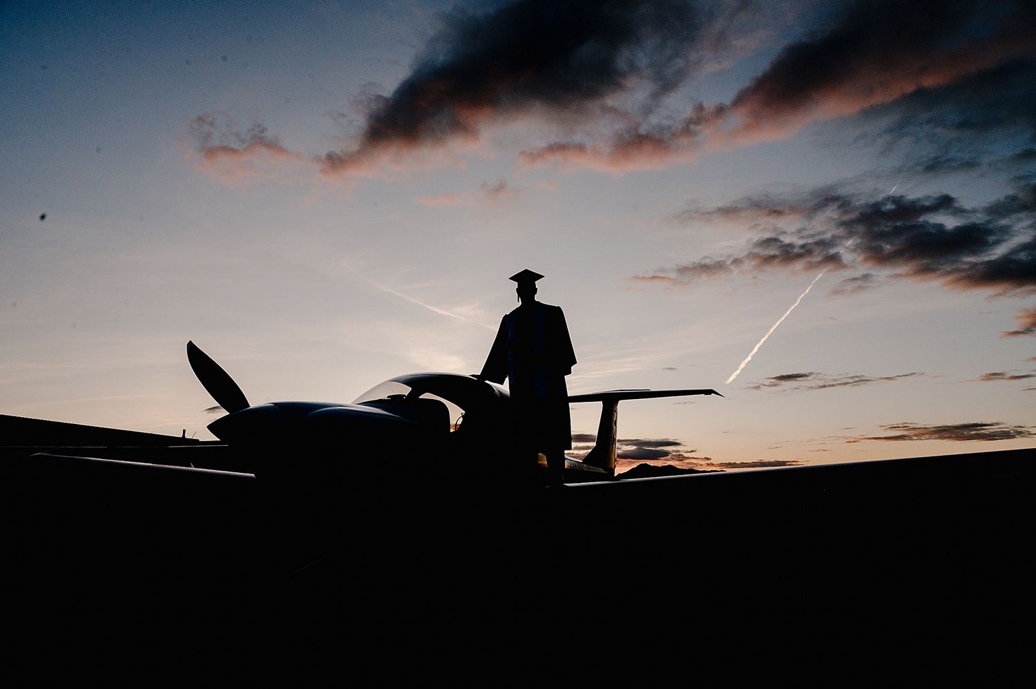 Silhouette figure standing beside small aircraft against dramatic sunset sky with streaking clouds.