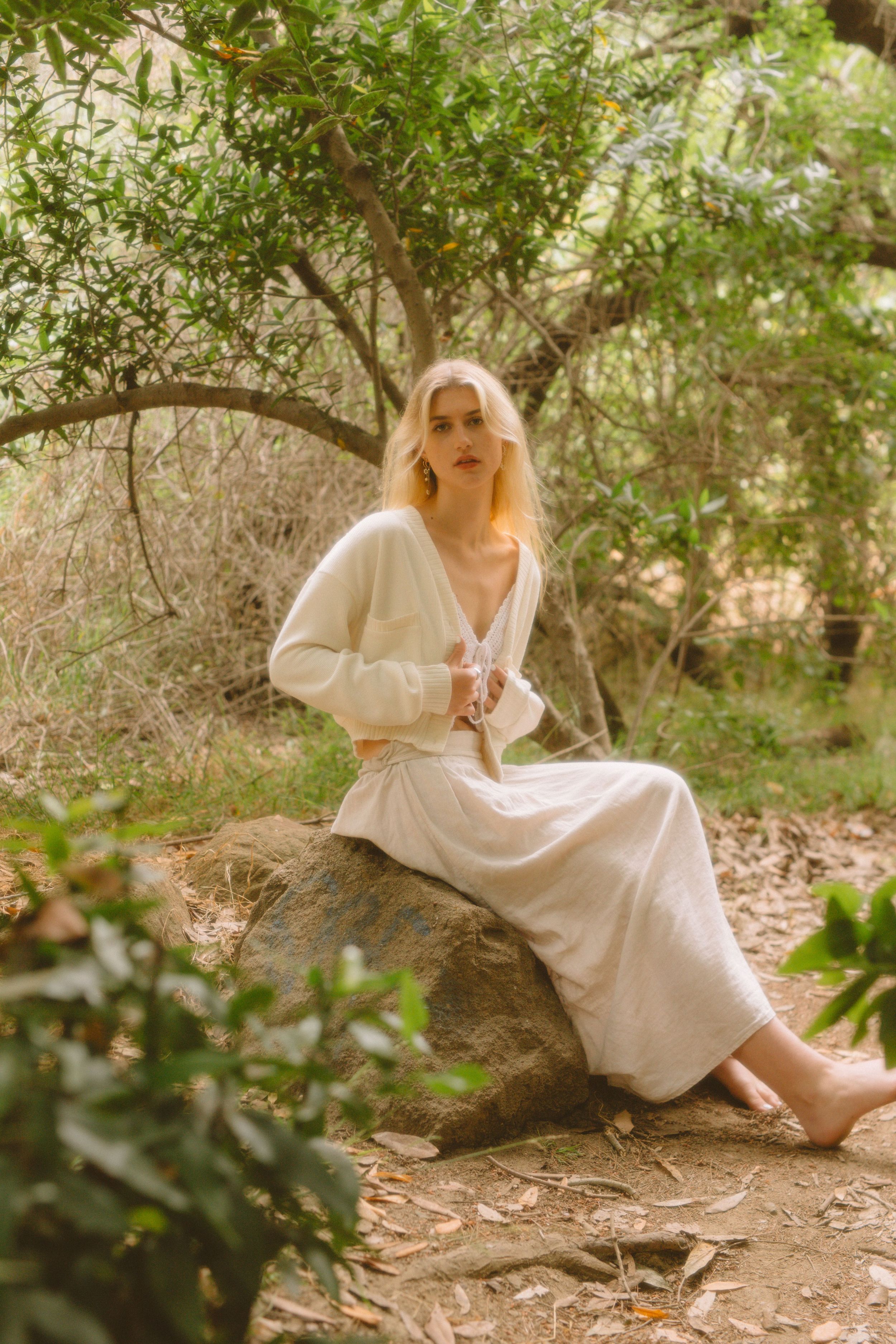Woman in white flowy dress sits on rock in ethereal garden setting with dappled sunlight.