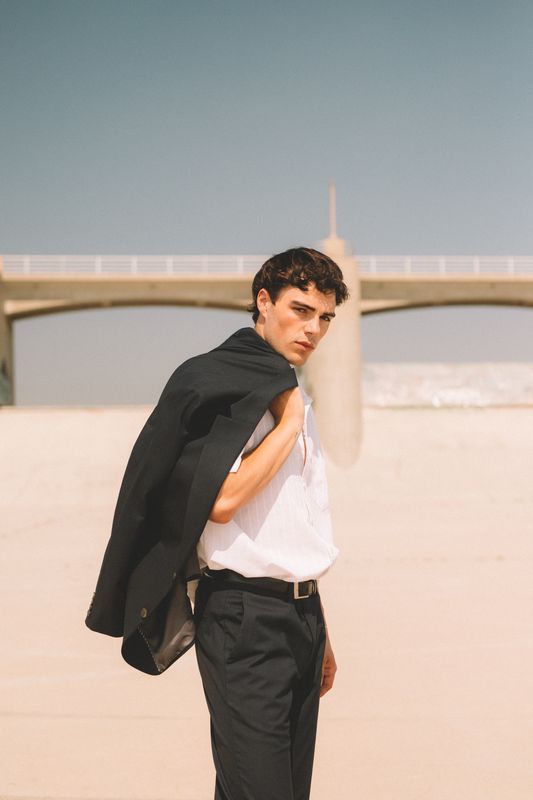 Dramatic fashion pose with black jacket and white shirt against architectural background.