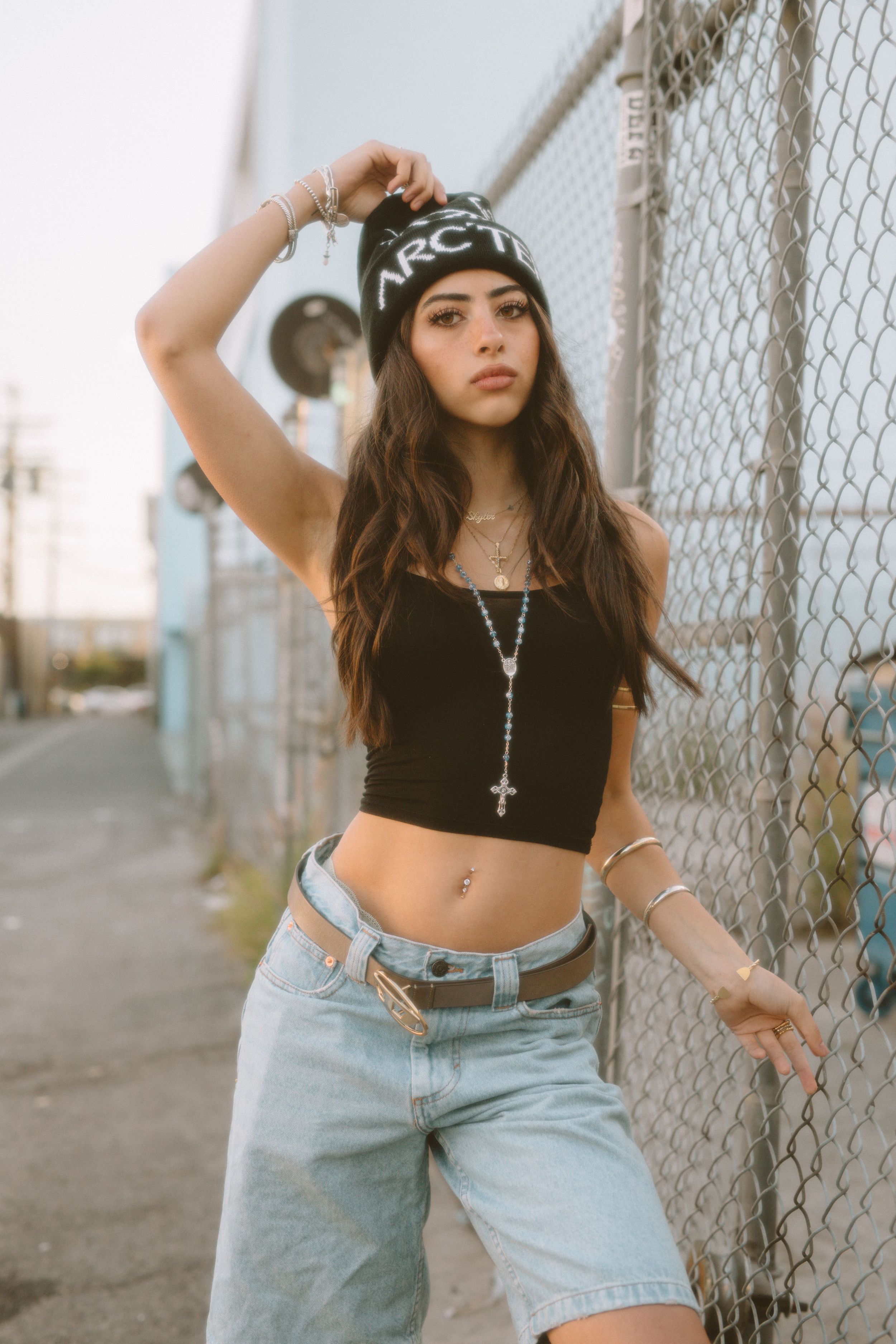 A fashion model in 90s-style denim shorts and beanie poses against an urban chain-link fence backdrop.