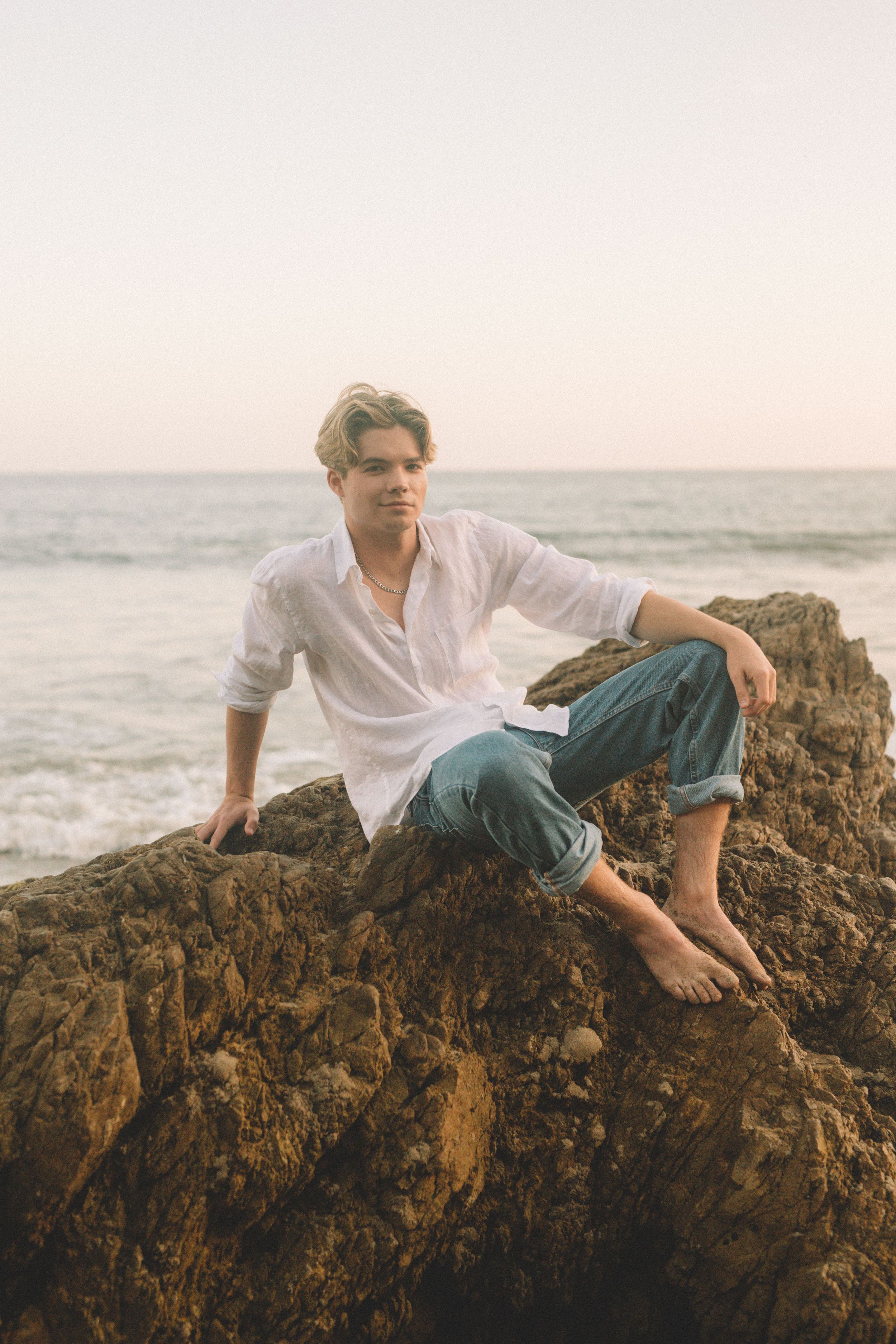 Person in white shirt and jeans poses on coastal rocks at sunset.