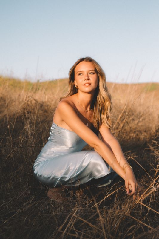 Woman in silver dress poses gracefully in tall grass during golden hour.
