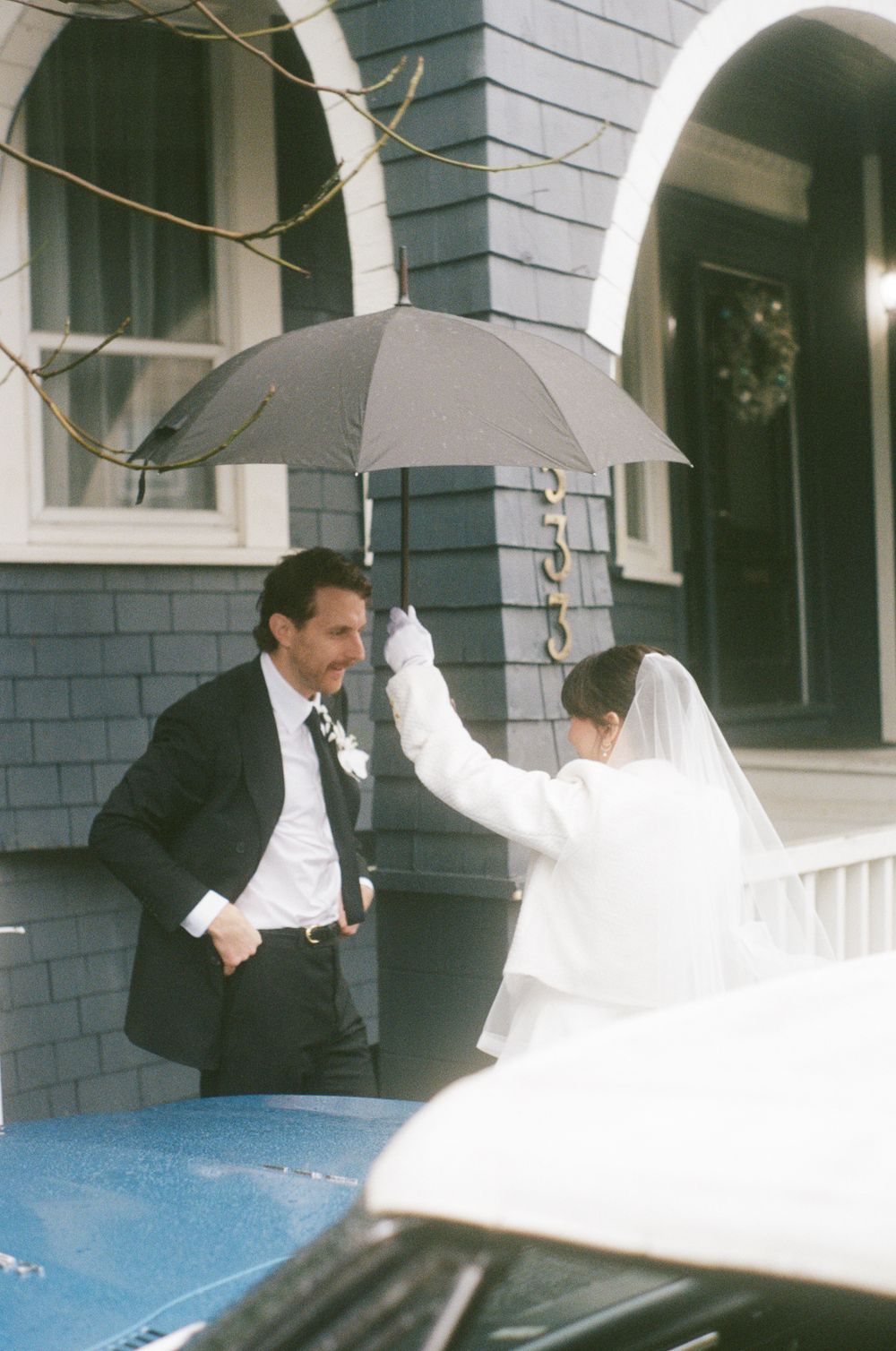 Wedding couple walking outside a grey house with white trim on a rainy day.