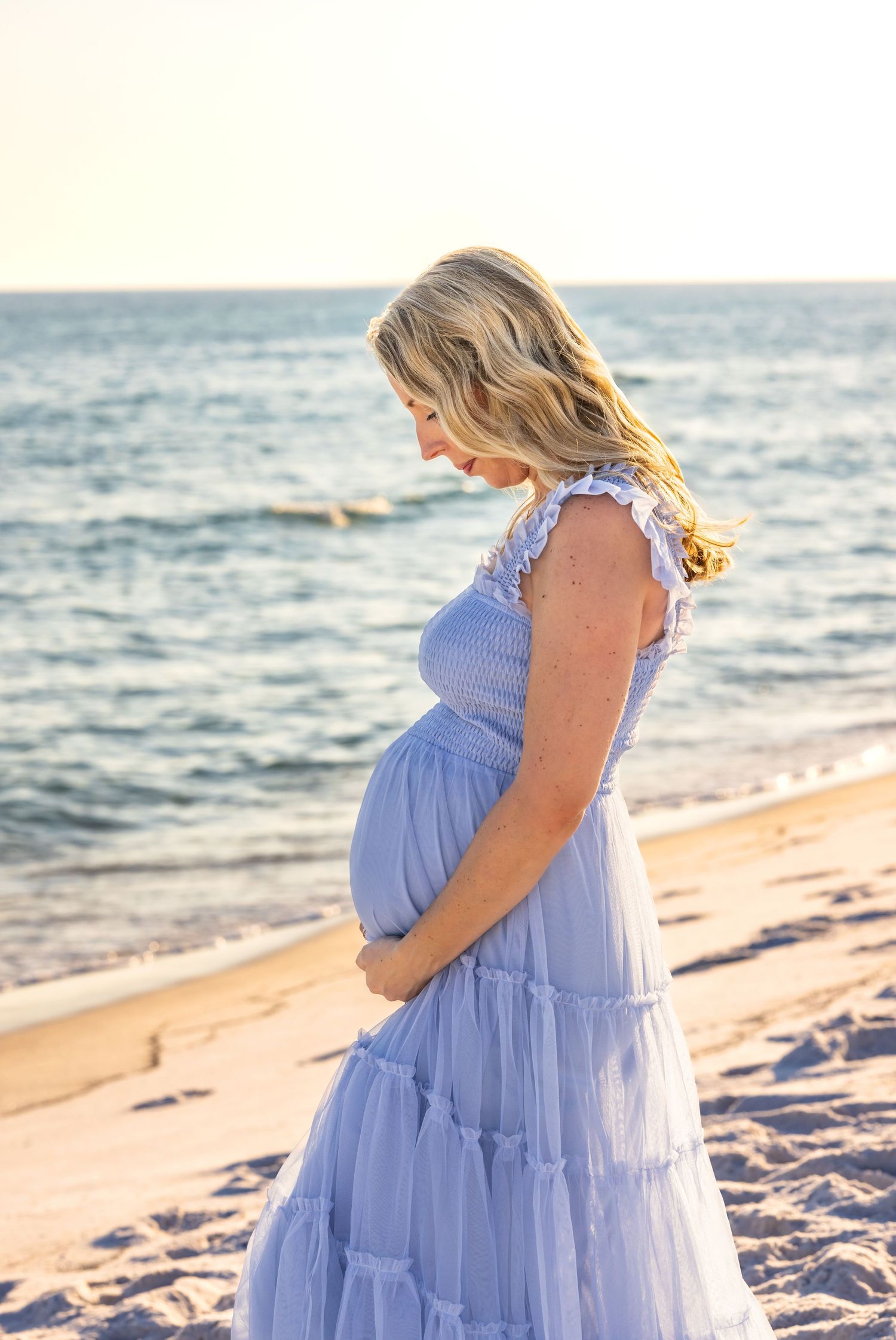 Someone in a flowing periwinkle dress stands on a sandy beach at sunset while cradling their pregnant belly.
