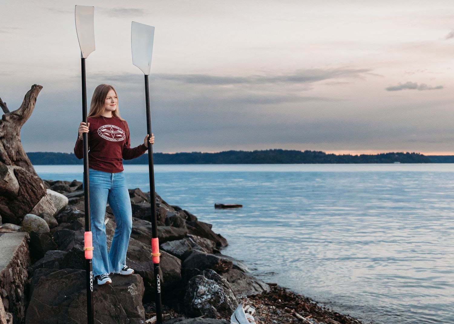 Adventurous paddler stands on rocky shoreline holding kayak oars against serene lake and cloudy sunset landscape.
