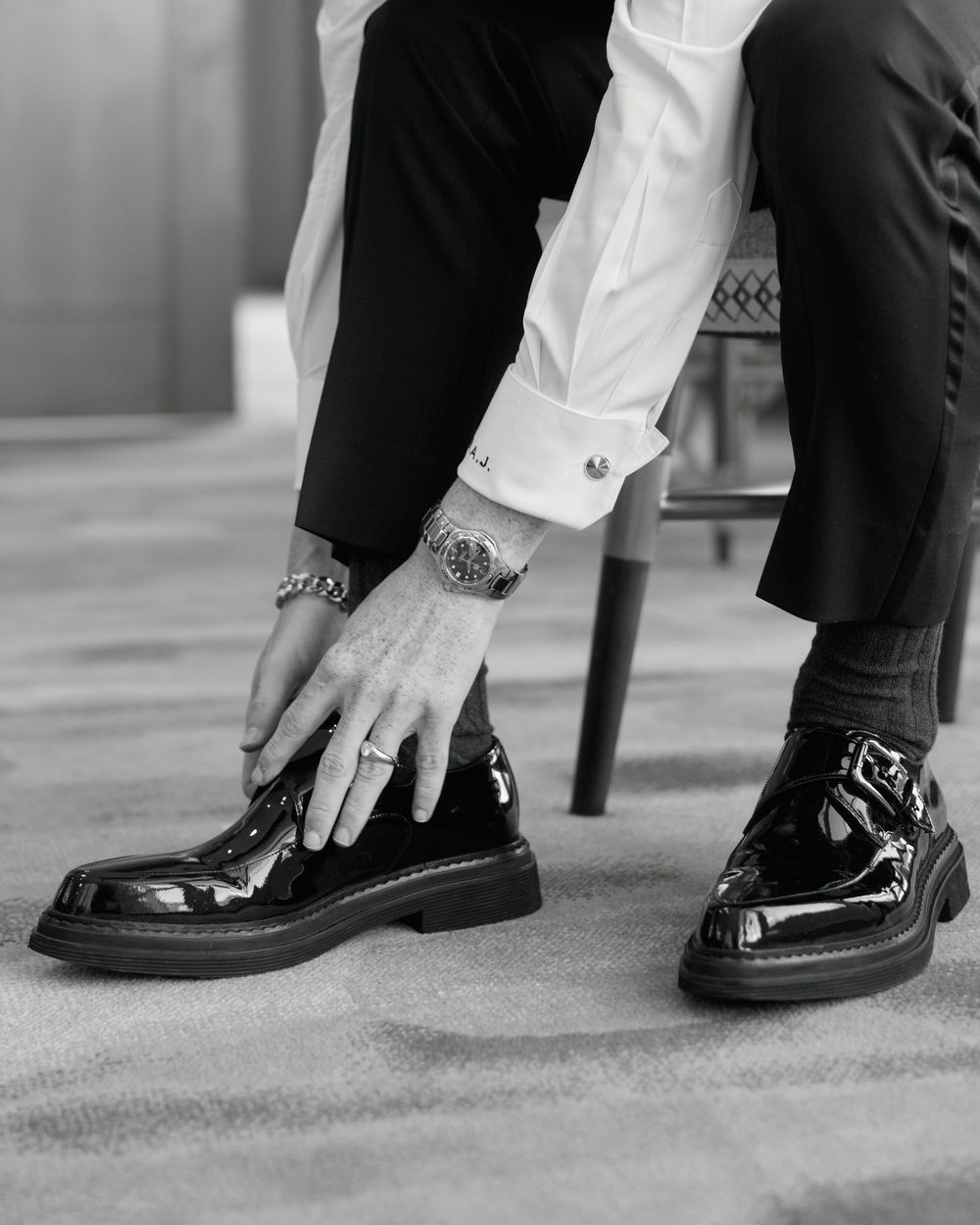 Black and white photo of hands adjusting patent leather dress shoes with jewelry and cufflinks visible.