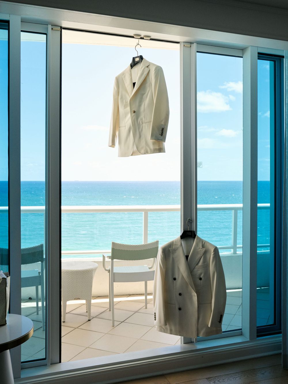 Two cream-colored suits hanging in a hotel room with ocean view through floor-to-ceiling windows.