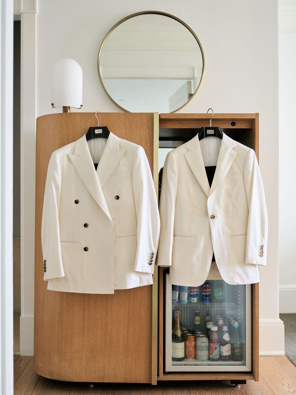 Two white formal suits hanging beside a round mirror and mini fridge in a hotel room.