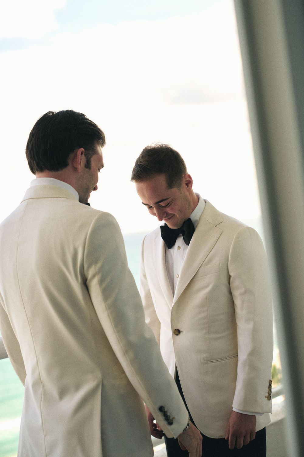 Two people in white tuxedo jackets having intimate moment on balcony with ocean view.