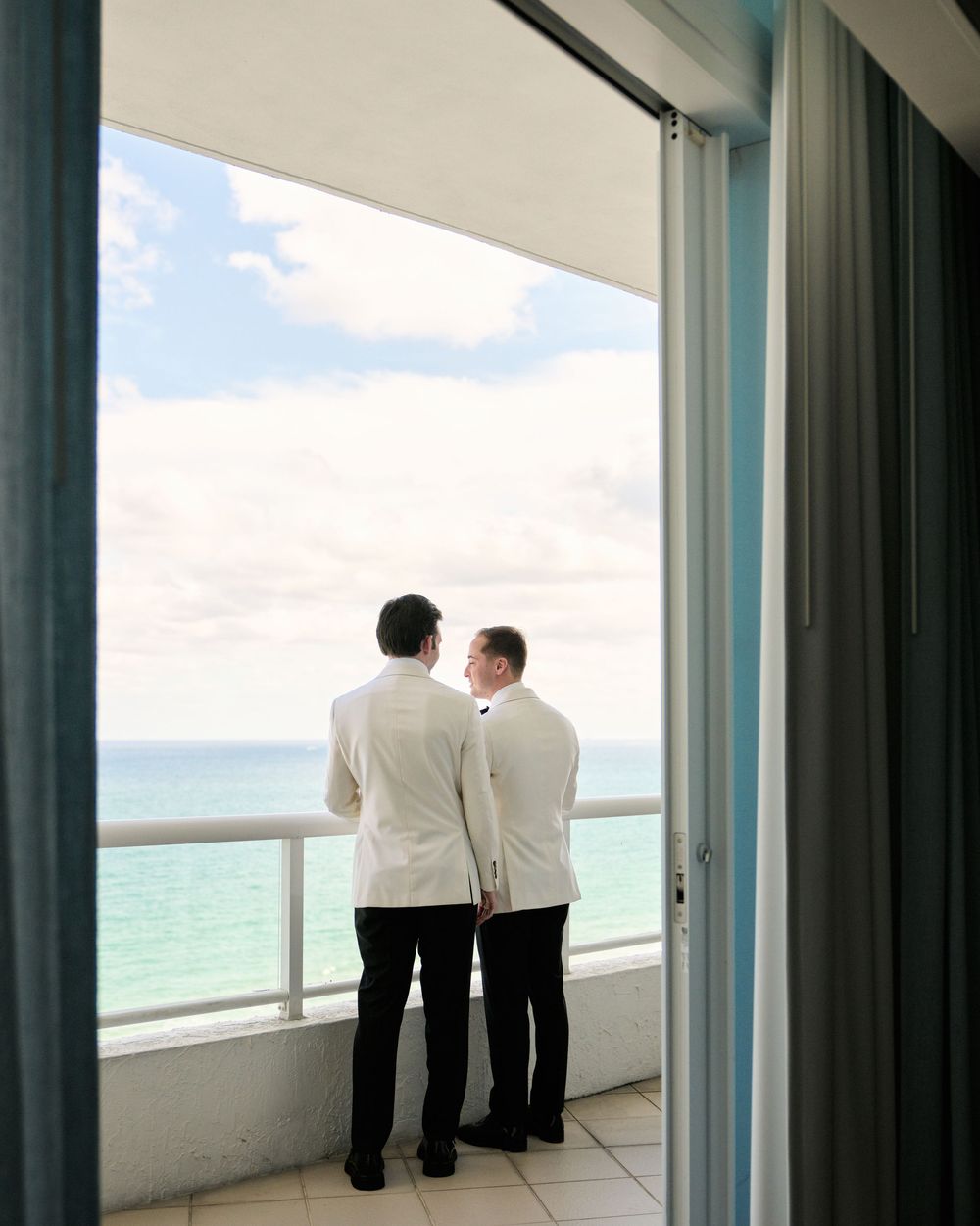 Two figures in white dinner jackets looking out over ocean from high-rise balcony.