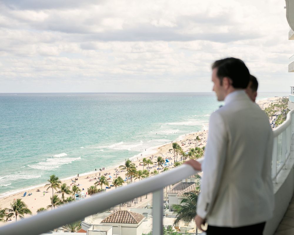A person in white looks out over a tropical beach view from a high balcony.