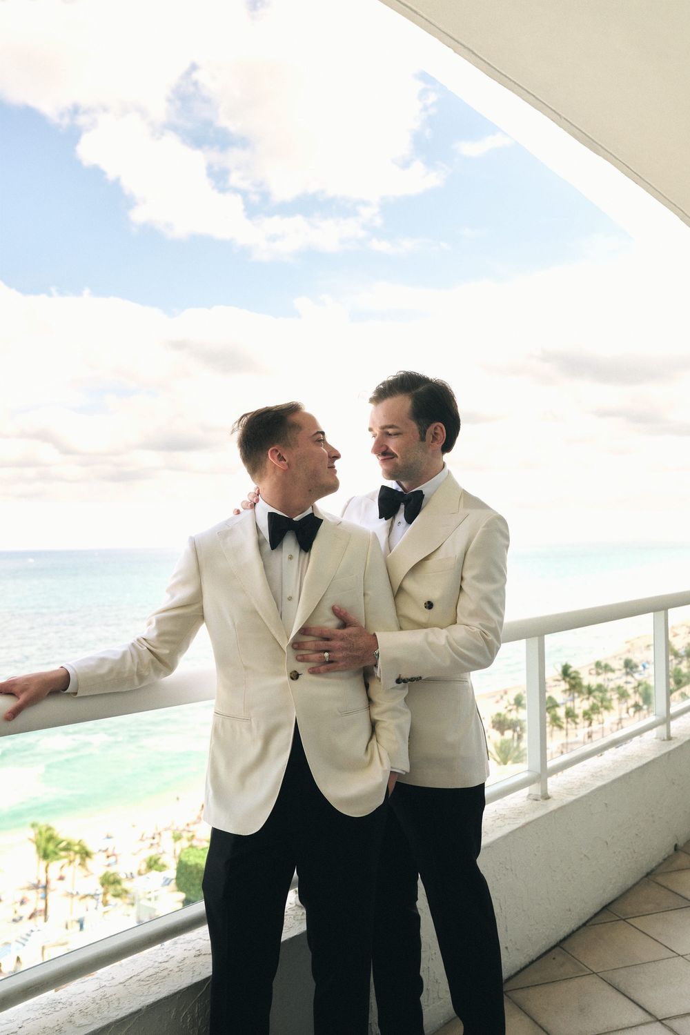 Two people in cream dinner jackets embrace on an oceanfront balcony.