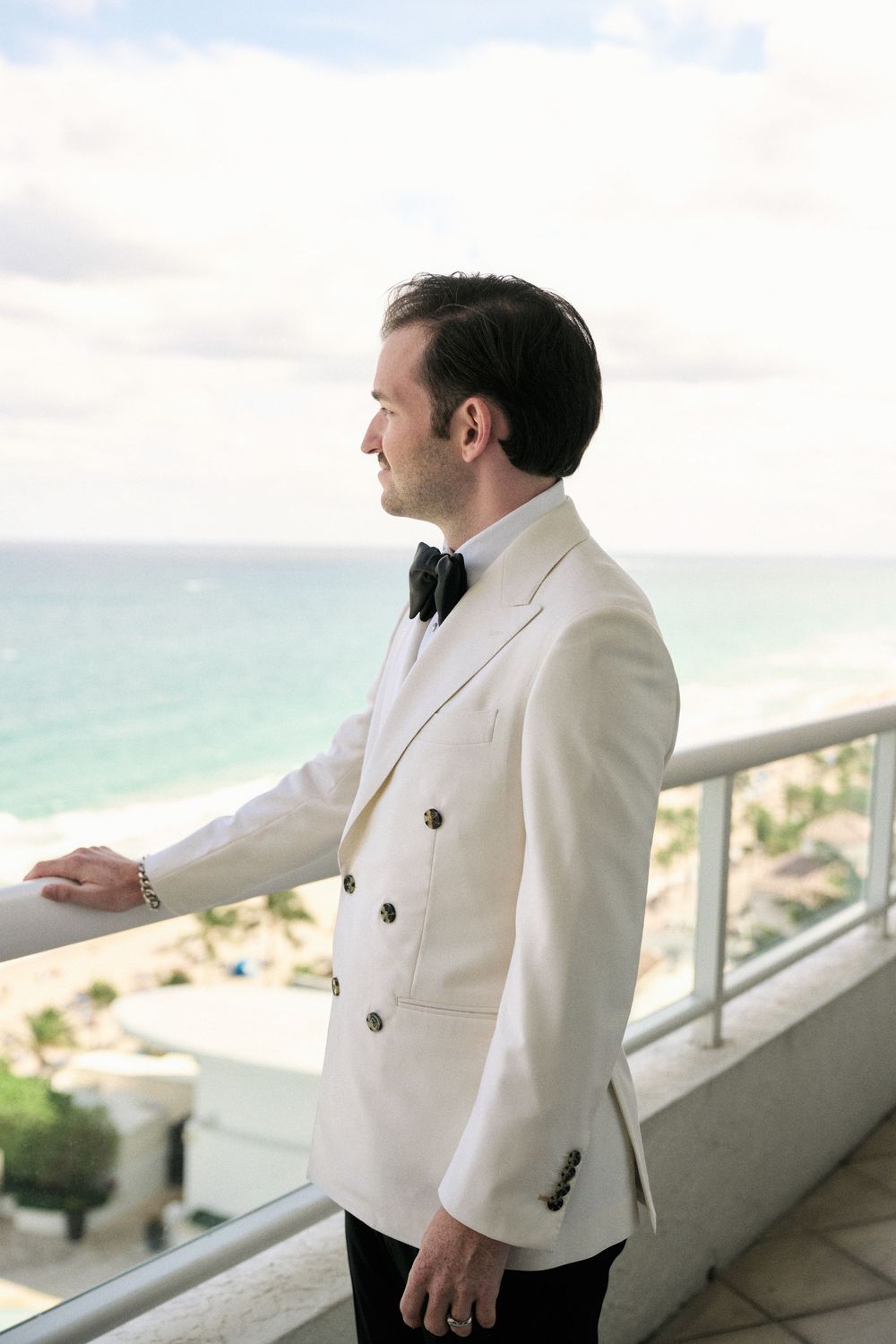 Person in white double-breasted tuxedo jacket standing on balcony overlooking tropical beach view.