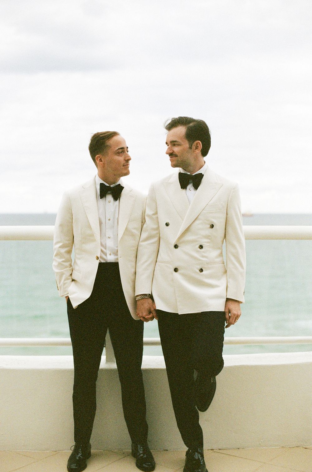 Two people in cream dinner jackets and bow ties stand together on an oceanfront balcony.