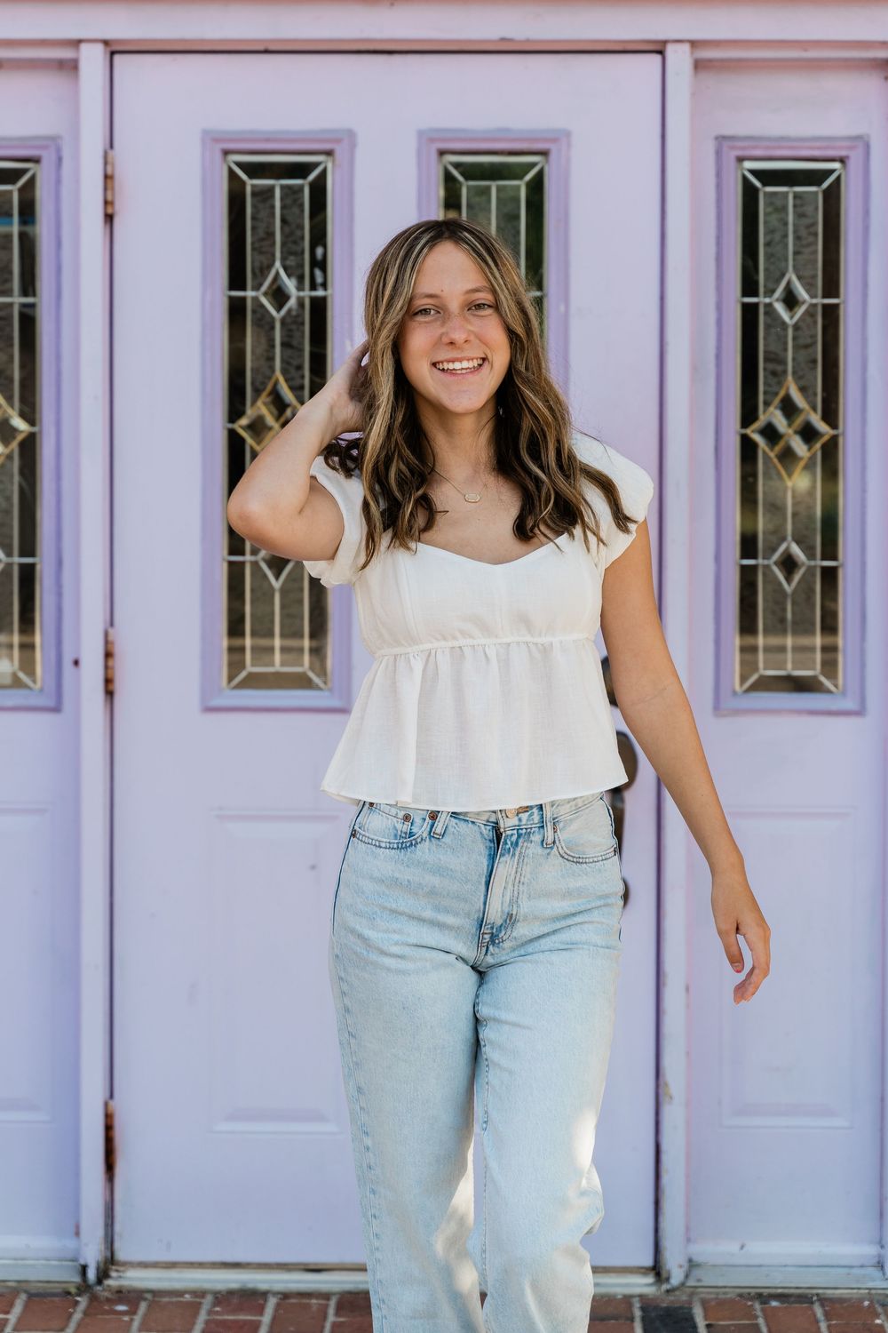 Woman in white babydoll top and light wash jeans poses casually in front of a lavender door with decorative glass panels.