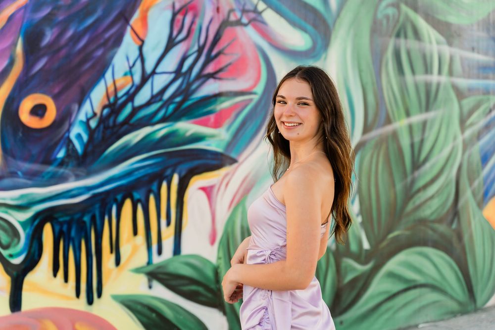 Smiling figure in lavender dress poses against vibrant tropical street art mural with peacock feather design.