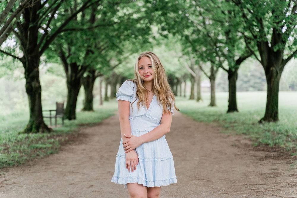 Someone in a light blue dress stands on a tree-lined dirt path in a park setting during summer.