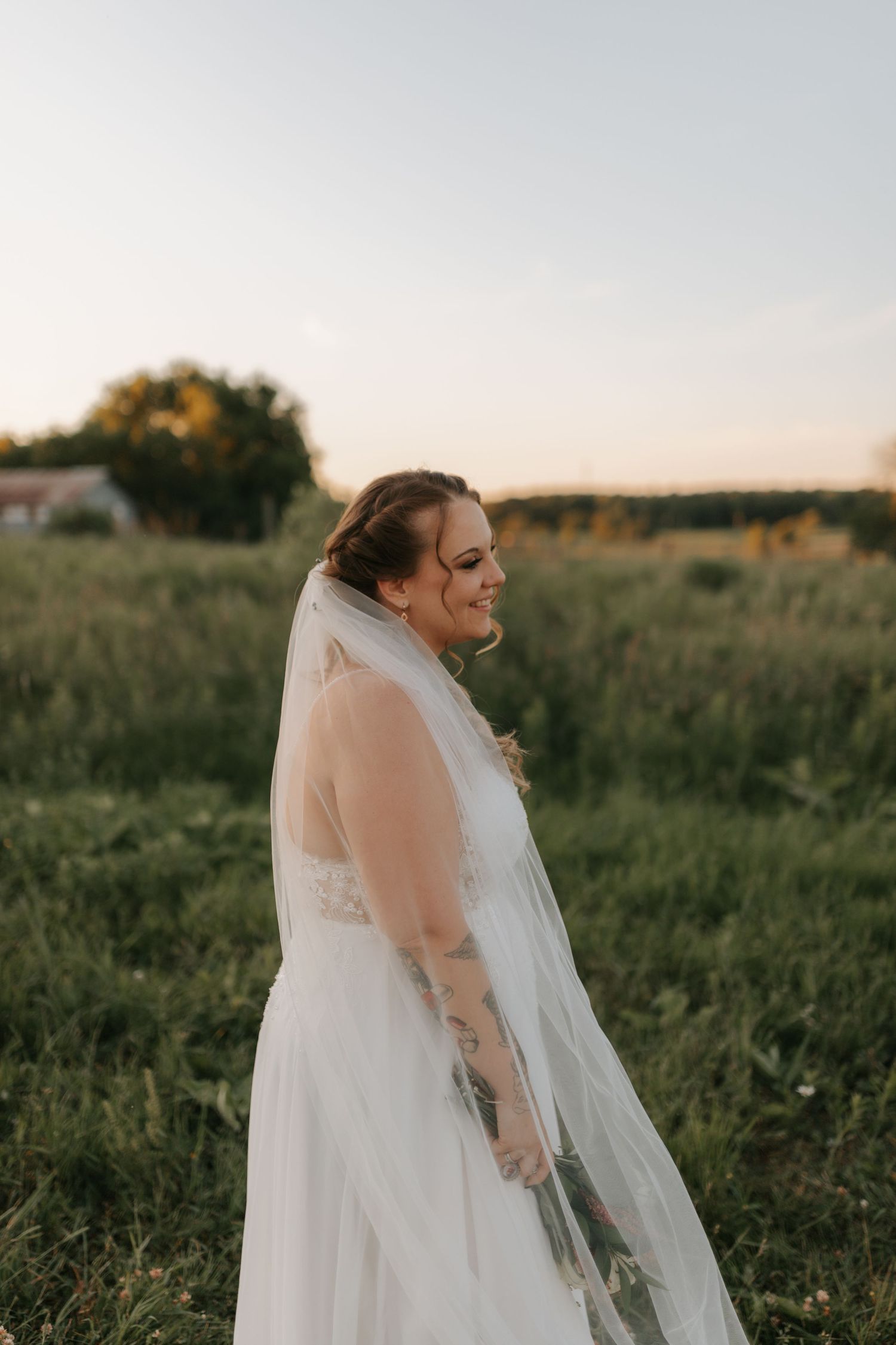 Bride in white gown and veil smiling in golden hour light in green field.
