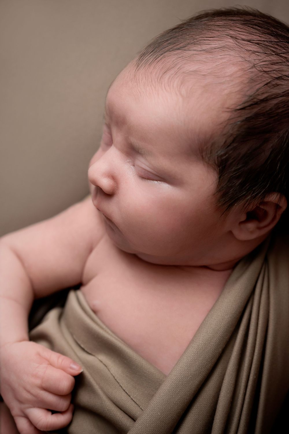 Close-up portrait of sleeping infant wrapped in tan cloth against brown background.