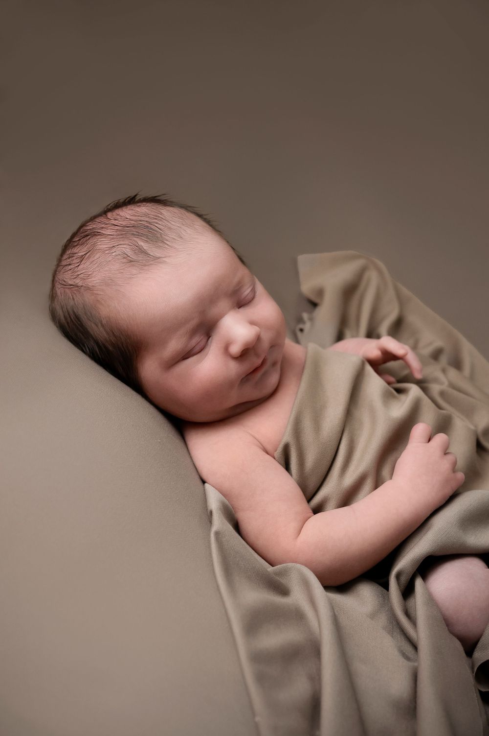 Peaceful sleeping newborn wrapped in soft tan blanket lying on neutral backdrop.
