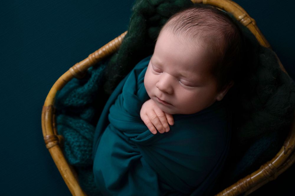 Peaceful newborn baby wrapped in teal blanket resting in bamboo basket against dark background.