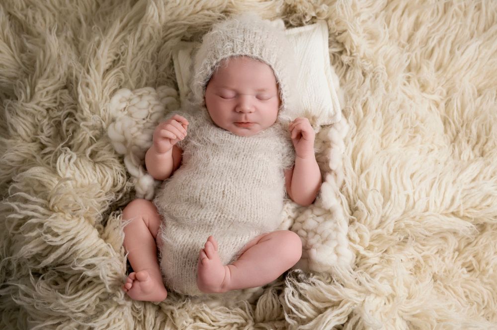 Newborn baby wearing white knit outfit lying on fluffy cream-colored rug.