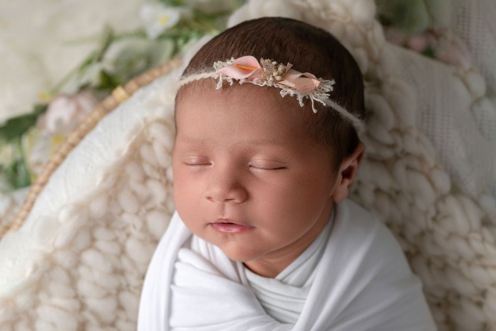 Sleeping newborn wearing white outfit and delicate floral headband on textured blanket.