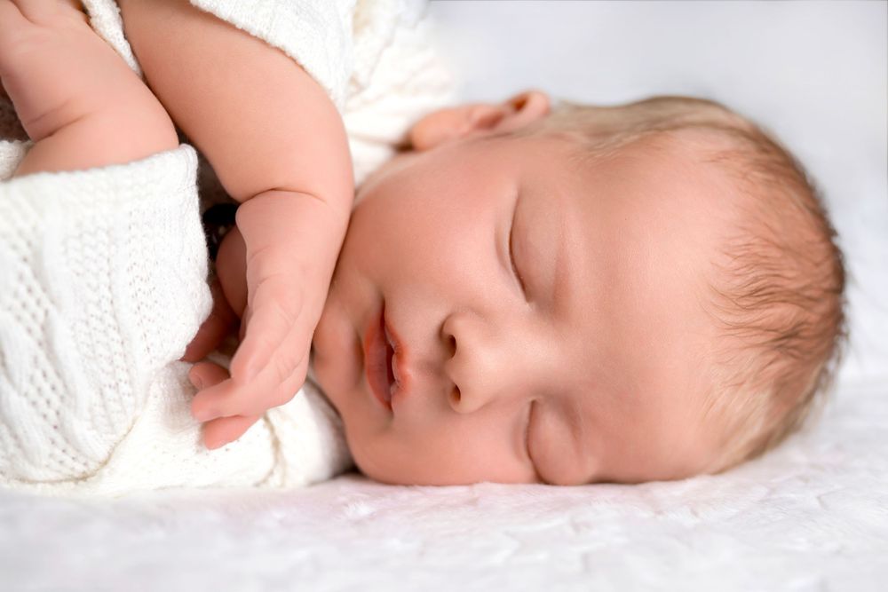 Sleeping baby in white knit outfit on soft white bedding.