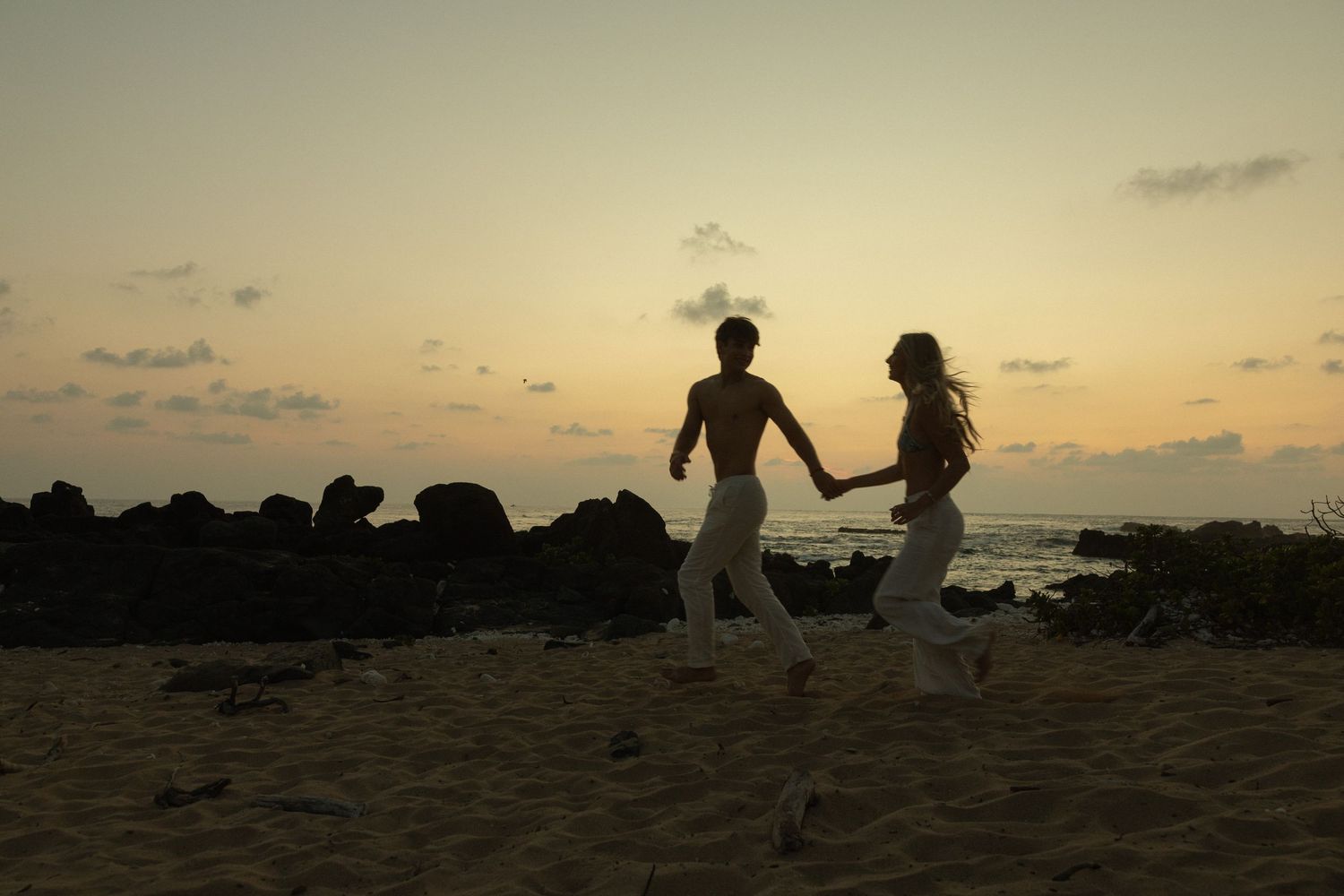 Two silhouettes dance playfully on a beach at sunset with rocky coastline and ocean waves in the background.