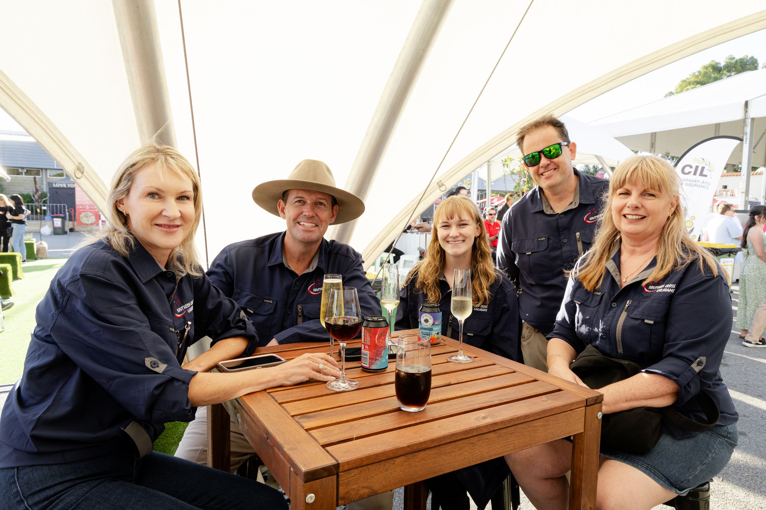 A group enjoying drinks and conversation at a wooden table under white canopy umbrellas at an outdoor event.