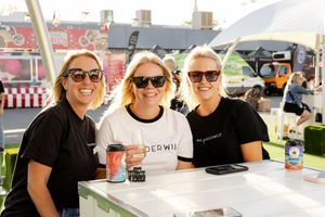 Three people in sunglasses sit together at an outdoor event booth while holding beverages and smiling.