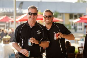 Two people in matching black polo shirts and sunglasses smiling and holding drinks at an outdoor event.