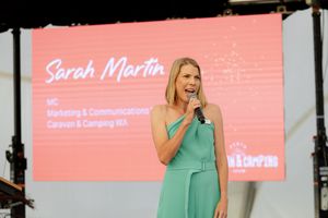 A presenter in a mint green dress speaks on stage with a pink presentation screen visible in the background.