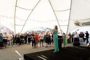 A crowd gathers under a white tent structure while someone speaks from an elevated stage during an outdoor event.
