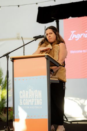 Speaker presents at Caravan and Camping podium during outdoor event with coral backdrop and string lights.