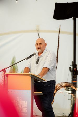 Speaker stands at a red podium on an outdoor stage wearing a white shirt during a public event.