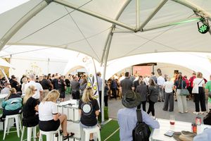 Large gathering of people under white canopy tent at outdoor social event.