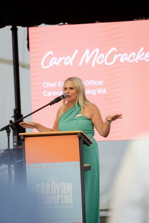 Presenter in green dress speaks at Caravan and Camping Western Australia podium with coral-colored backdrop.