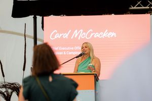 A speaker presents at a podium on stage with a pink backdrop screen displaying text.