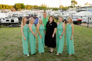 Group wearing matching seafoam green jumpsuits poses together at a marina with RVs in the background.