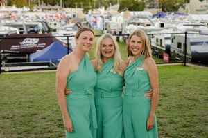 Three attendees in matching seafoam green dresses pose together on grass at a marina event.